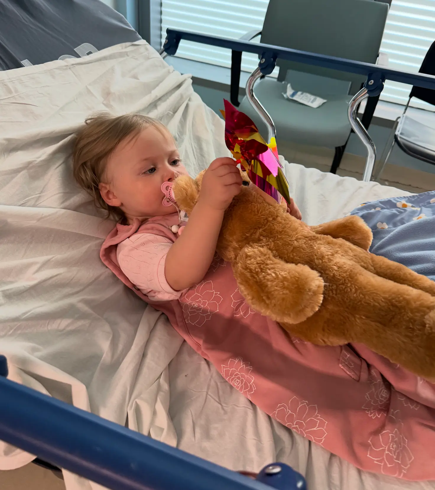 A toddler resting in a hospital bed holding a teddy bear